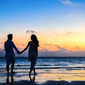 A romantic couple walks hand in hand on a tropical beach at sunset, enjoying a serene moment together.