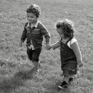 Two young children joyfully holding hands and walking in a grassy field, captured in charming black and white.